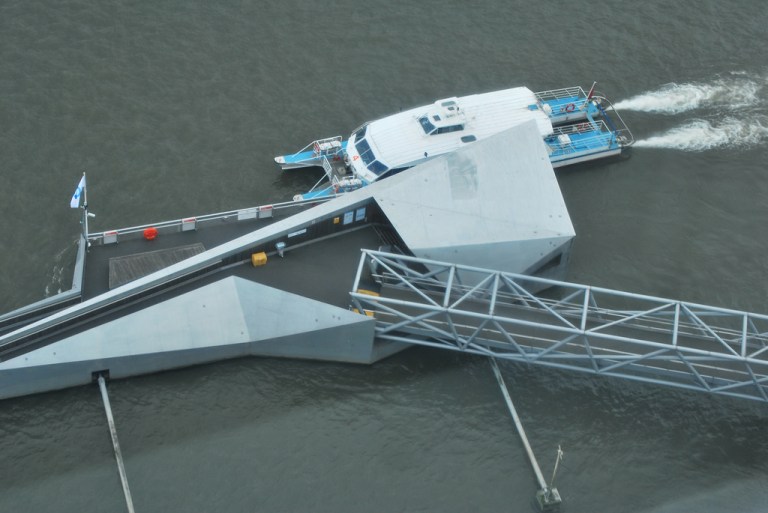 Millbank Millennium Pier from above. The ramp descends in two sections to the left of the point at which the brow meets the pier, while the stairs are under cover on the right. By distillated [CC  BY-SA 2.0] via this flickr page